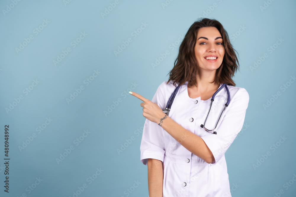 Young beautiful woman doctor in a lab coat on a blue background smiles, and points her finger to the left on an empty space Young beautiful woman doctor in a lab coat on a blue background smiles, and points her finger to the left on an empty space