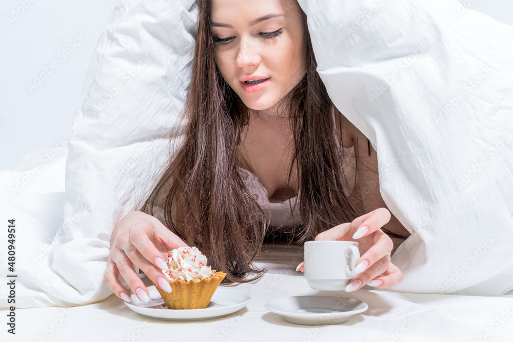 A woman sits in bed and drinks aromatic coffee and eats a delicious cupcake. Tasty breakfast. The brunette holds a cup with a hot drink and an appetizing cupcake. White bedroom. Good morning