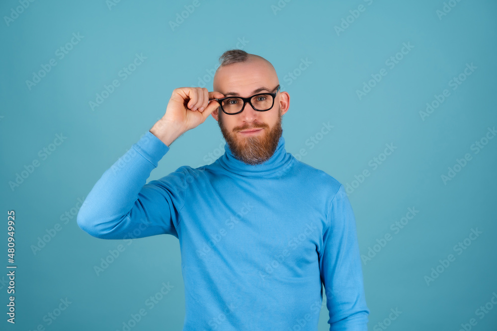 A young man with a red beard in a turtleneck and glasses on a blue background looks at the camera with a confident smile A young man with a red beard in a turtleneck and glasses on a blue background looks at the camera with a confident smile