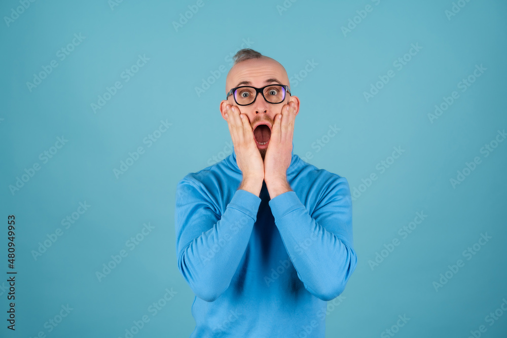 A young man with a red beard in a turtleneck and glasses on a blue background looks at the camera, surprised shocked with his mouth open A young man with a red beard in a turtleneck and glasses on a blue background looks at the camera, surprised shocked with his mouth open