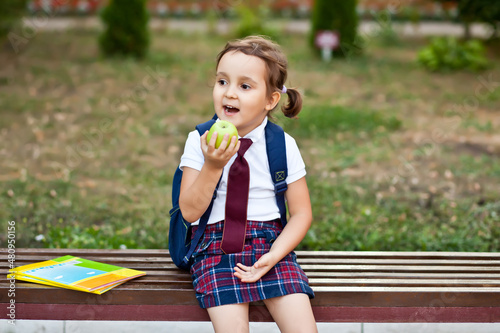 little cute schoolgirl in uniform sitting on a bench and eating an apple
