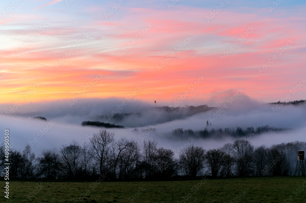 Sauerland scenery panorama on a foggy winters evening after sunset. Colorful sky and wafts of mist and clouds in the valleys near Iserlohn and Altena Germany. Mystic atmosphere in rural landscape.