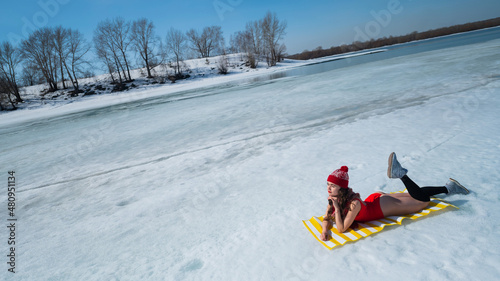 Caucasian woman in a red swimsuit and a knitted hat sunbathes in winter lying on the snow.
