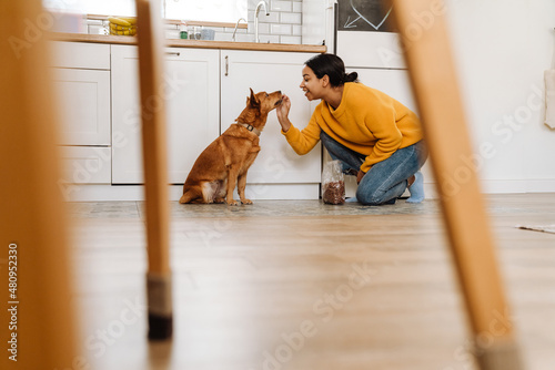 Young hispanic woman smiling while feeding her dog in kitchen