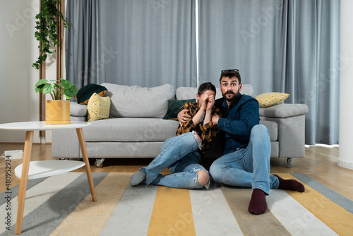 Young couple sitting on the floor watching horror movie woman covered her face from fear and man making terrified facial expression as he is scared. 