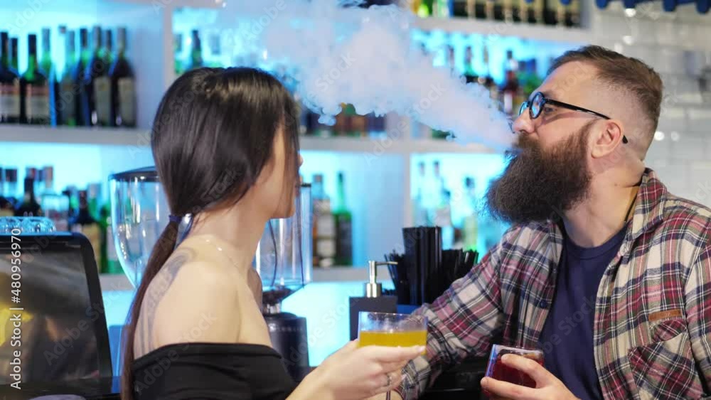 A couple enjoys a date, talking and drinking alcoholic cocktails together in a stylish nightclub.A brutal bearded man smokes an electronic cigarette behind the bar.The concept of rest and love A couple enjoys a date, talking and drinking alcoholic cocktails together in a stylish nightclub.A brutal bearded man smokes an electronic cigarette behind the bar.The concept of rest and love