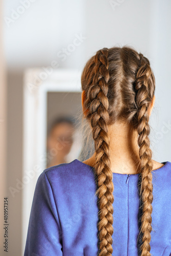 a person with long hair looks at himself in the mirror with two braids. actual color dress. healthy beautiful hair.