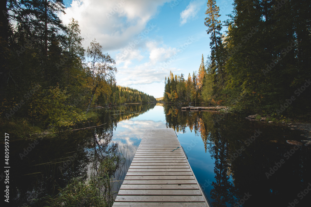 View of a wooden pier and a still river reflecting the colourful autumn forest with all its colours. Wonderful autumn in the barren wilderness of Hossa National Park, in Lapland, northern Finland View of a wooden pier and a still river reflecting the colourful autumn forest with all its colours. Wonderful autumn in the barren wilderness of Hossa National Park, in Lapland, northern Finland