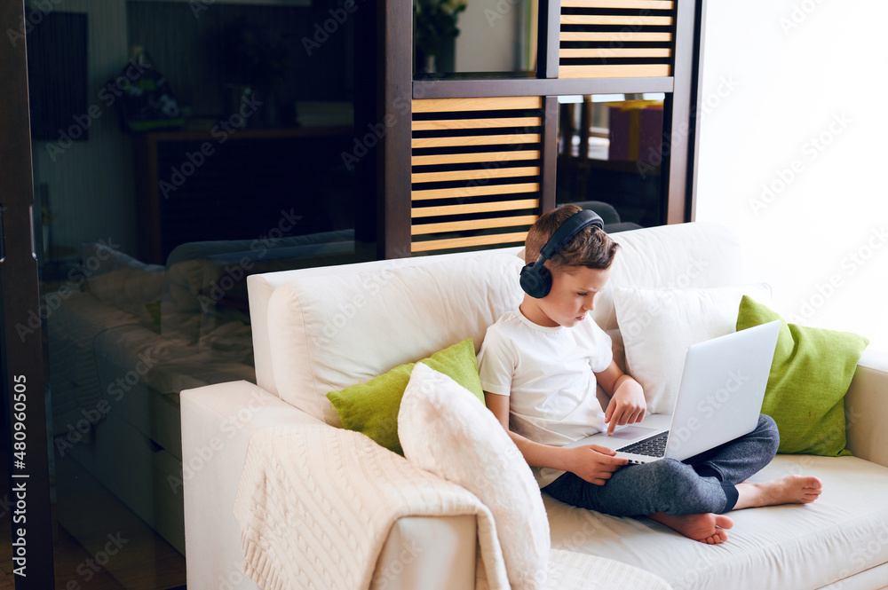 Concentrated schoolboy wearing headphones with laptop play online games at home. Distance learning in quarantine