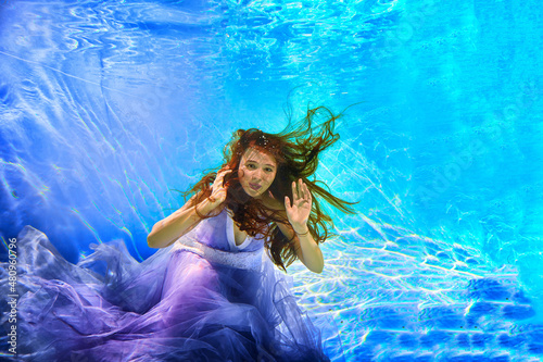 Girl in a beautiful dress under blue water. Female model posing under water in swimming pool