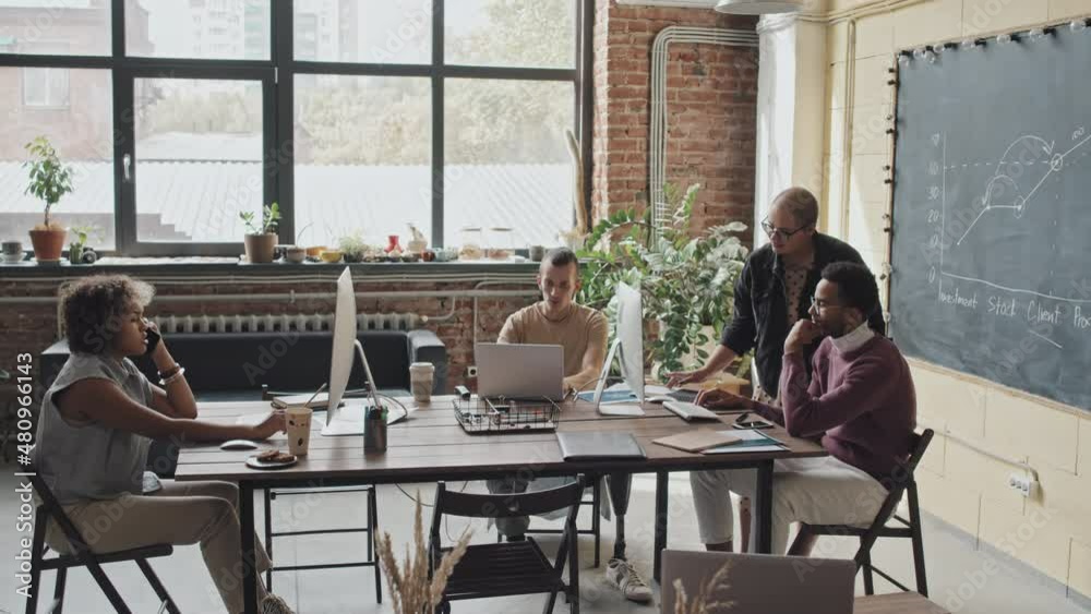 Long shot of four multiethnic colleagues using computers, sitting at table in loft-styled coworking space at daytime, working on project Long shot of four multiethnic colleagues using computers, sitting at table in loft-styled coworking space at daytime, working on project