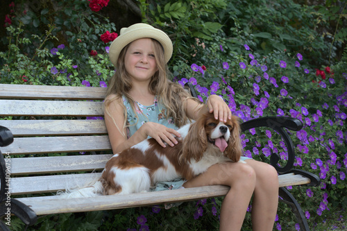 Blonde girl in a straw hat sits on a bench with a dog King Charles Cavalier Spaniel