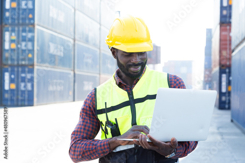 Portrait Black American African warehouse staff wearing yellow hard hat, construction vest and ear protector standing with happy smile and checking list of container boxes for shipping logistic stock.