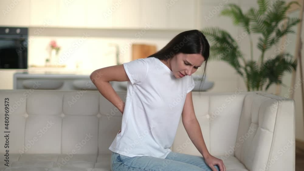 Back spasm. Upset frustrated caucasian young adult woman suffering from backache, unhappy brunette sit on a sofa in living room, feels discomfort because of pain in back, need treatment and rest
