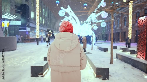 rear view camera following woman in knitted red cap and scarf walking on city square with festive colorful illuminations at night. soft snow is falling and snowflakes flying around.
