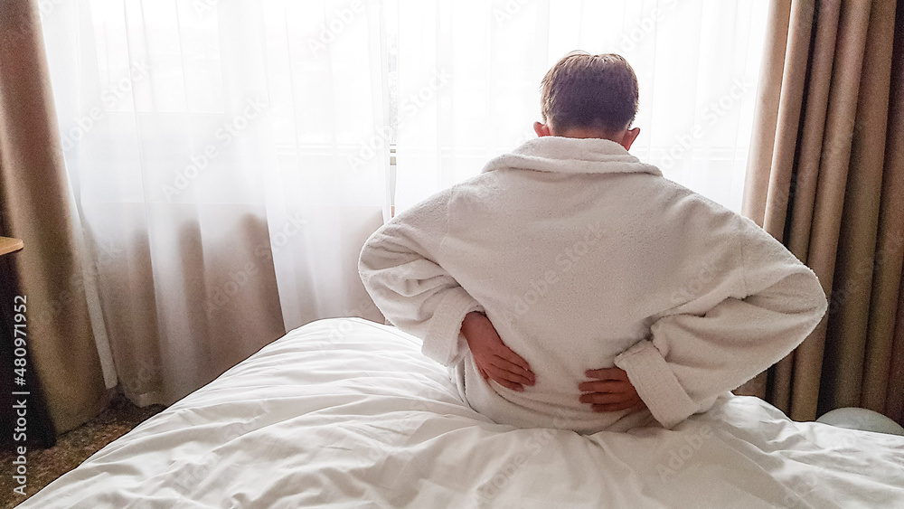 a young man with blond hair is sitting on the bed in a white robe and holding his hands behind his back. my back hurts. get up early in the morning