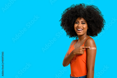 Fotografie, Obraz Black teen girl smile and points to her arm with vaccine sticker, she does not wear face shield, isolated on blue background