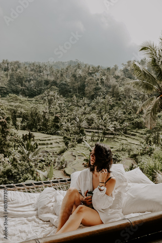 A young travelling woman relaxing in the lounge area of a Bali jungle hotel surrounded by jungle, palm trees and nature