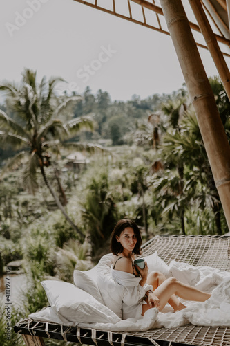 A young travelling woman relaxing in the lounge area of a Bali jungle hotel surrounded by jungle, palm trees and nature