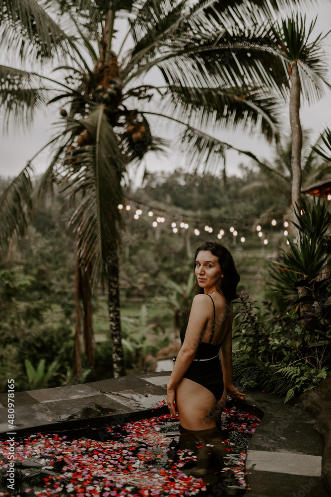 Fototapeta premium A young female traveller is relaxing by the small plunge pool of a jungle hotel in Bali surrounded by jungle, rice fields and tropical landscape