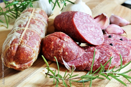 Traditional smoked salami sausages sliced with pepper, garlic and rosemary on wooden cutting board.