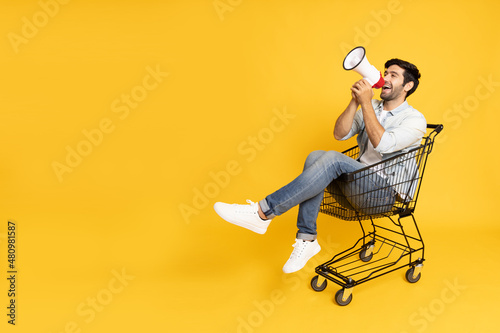 Young man sitting inside of shopping trolley and holding megaphone  isolated on yellow background, Announce concept