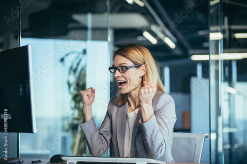 Beautiful and successful business woman with glasses, celebrating victory looks at the computer monitor screen and rejoices in the successfully completed project, keeps her hands up from 