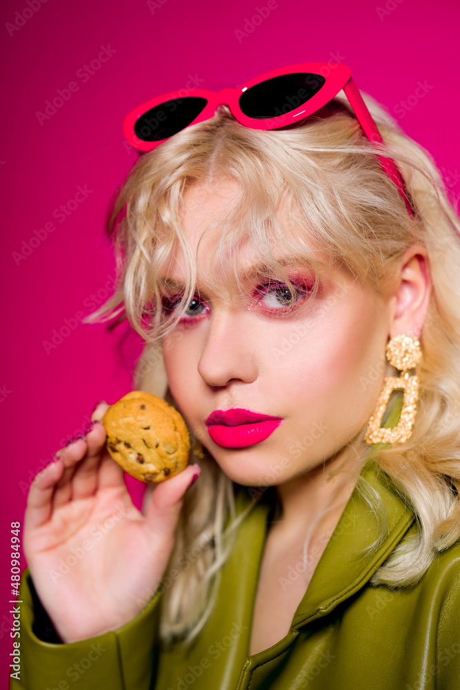 Obraz premium The blonde is eating cookies. High fashion, woman in earrings, gold jewelry. Bright pink glitter makeup. Pink glasses and stylish green cape