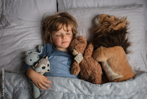 Healthy child, sweetest blonde toddler boy sleeping in a bed with a teddy bear and another stuffed animals. Beautiful baby boy have a healthy sleep in the bed. Sleeping baby.