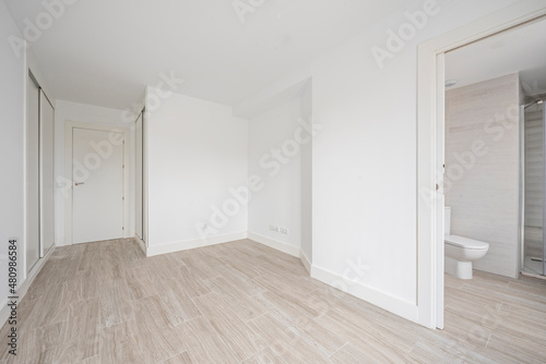 Empty room with white fitted wardrobes, white walls and wood-like ceramic tile floors and en-suite bathroom