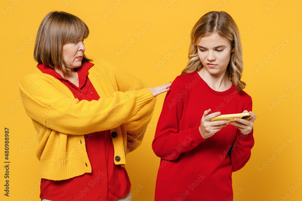 Strict woman 50s wear red shirt with girl 12-13 years old. Grandmother granddaughter use play racing app on mobile cell phone hold gadget smartphone pc video games isolated on plain yellow background Strict woman 50s wear red shirt with girl 12-13 years old. Grandmother granddaughter use play racing app on mobile cell phone hold gadget smartphone pc video games isolated on plain yellow background