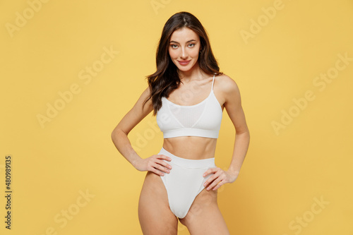 Smiling skinny sexy lovely attractive young brunette woman 20s wearing white underwear with beautiful perfect fit body standing akimbo posing looking camera isolated on plain yellow background studio