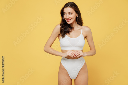 Smiling minded attractive young brunette woman 20s in white underwear with perfect fit body standing posing look aside on workspaece area mock up isolated on plain yellow background studio portrait