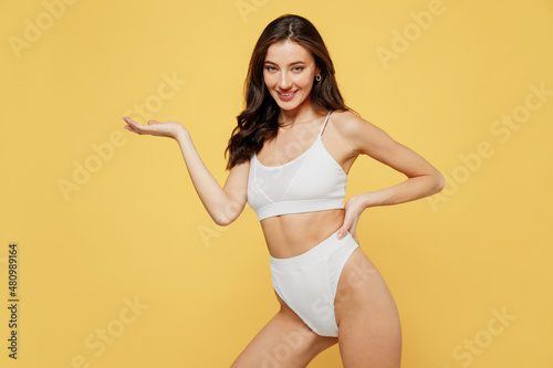 Smiling lovely attractive young brunette woman 20s in white underwear with perfect fit body standing posing hold show empty arm with workspace area isolated on plain yellow background studio portrait © ViDi Studio