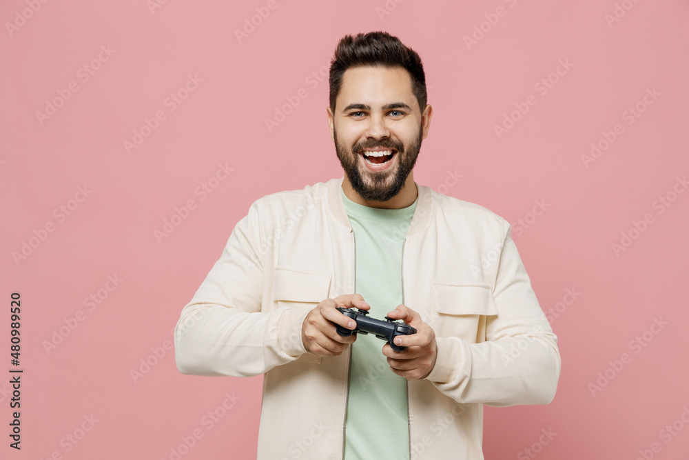 Young smiling happy caucasian man 20s wear trendy jacket shirt hold in hand play pc game with joystick console isolated on plain pastel light pink background studio portrait. People lifestyle concept. Young smiling happy caucasian man 20s wear trendy jacket shirt hold in hand play pc game with joystick console isolated on plain pastel light pink background studio portrait. People lifestyle concept.