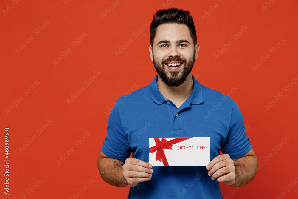 Young smiling happy fun man 20s wear basic blue t-shirt looking camera hold gift certificate coupon voucher card for store isolated on plain orange background studio portrait People lifestyle concept Young smiling happy fun man 20s wear basic blue t-shirt looking camera hold gift certificate coupon voucher card for store isolated on plain orange background studio portrait People lifestyle concept