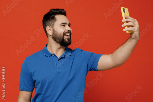 Young smiling happy man 20s wear basic blue t-shirt looking camera doing selfie shot on mobile cell phone post photo on social network isolated on plain orange background. People lifestyle concept