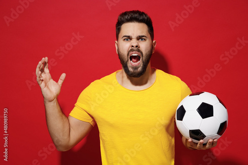 Screaming scolding upset angry young bearded man football fan in yellow t-shirt cheer up support favorite team hold soccer ball yell raised hand isolated on plain dark red background studio portrait.
