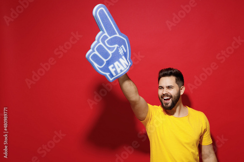 Smiling excited overjoyed young bearded man 20s in yellow t-shirt cheer up support favorite sport team look aside hold fan foam glove finger up isolated on plain dark red background studio portrait. © ViDi Studio
