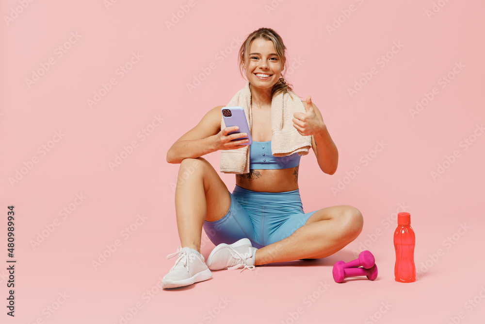 Full size young fitness trainer instructor woman wear blue tracksuit spend time in home gym sit on floor use mobile cell phone show thumb up isolated on plain pink background. Workout sport concept Full size young fitness trainer instructor woman wear blue tracksuit spend time in home gym sit on floor use mobile cell phone show thumb up isolated on plain pink background. Workout sport concept