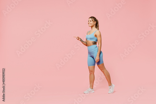 Full body fun young strong sporty fitness trainer woman wear blue tracksuit spend time in home gym walk go point finger aside on area isolated on pastel plain pink background. Workout sport concept.
