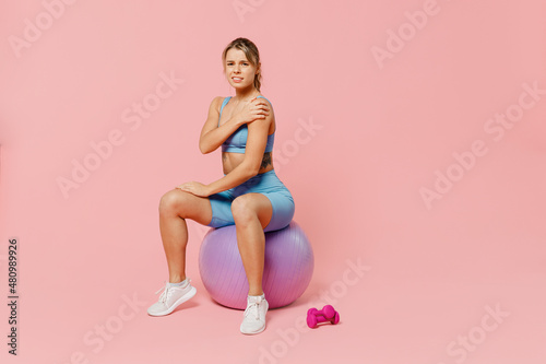 Full body young sporty fitness trainer instructor woman wear blue tracksuit spend time in home gym sit on fitball hold hand suffer from pain isolated on plain pink background. Workout sport concept.