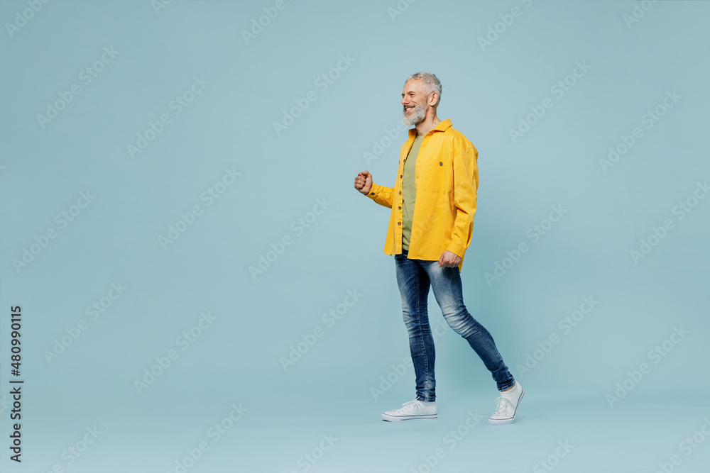 Full body cheerful happy elderly gray-haired mustache bearded man 50s wear yellow shirt walk going strolling isolated on plain pastel light blue background studio portrait. People lifestyle concept.
