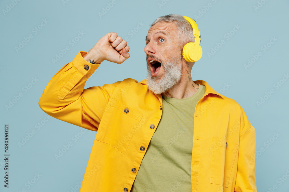 Elderly happy gray-haired mustache bearded man 50s wear yellow shirt headohones listen to music dance sing song in microphone isolated on plain pastel light blue background. People lifestyle concept. Elderly happy gray-haired mustache bearded man 50s wear yellow shirt headohones listen to music dance sing song in microphone isolated on plain pastel light blue background. People lifestyle concept.