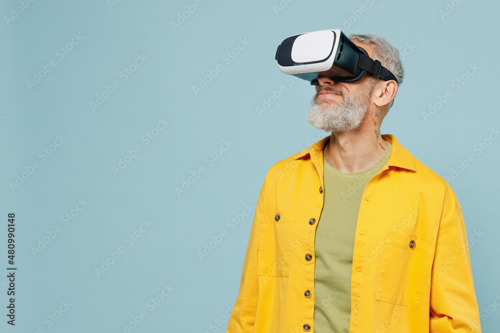 Elderly amazed happy gray-haired mustache bearded man 50s wear yellow shirt watching in vr headset pc gadget isolated on plain pastel light blue background studio portrait. People lifestyle concept. Elderly amazed happy gray-haired mustache bearded man 50s wear yellow shirt watching in vr headset pc gadget isolated on plain pastel light blue background studio portrait. People lifestyle concept.