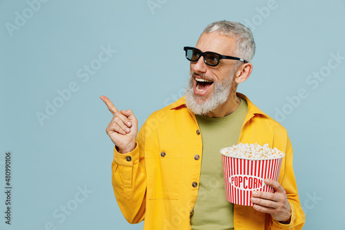 Young elderly man in 3d glasses watch movie film hold bucket of popcorn point finger aside on workspace isolated on plain blue background studio portrait. People emotions in cinema lifestyle concept.