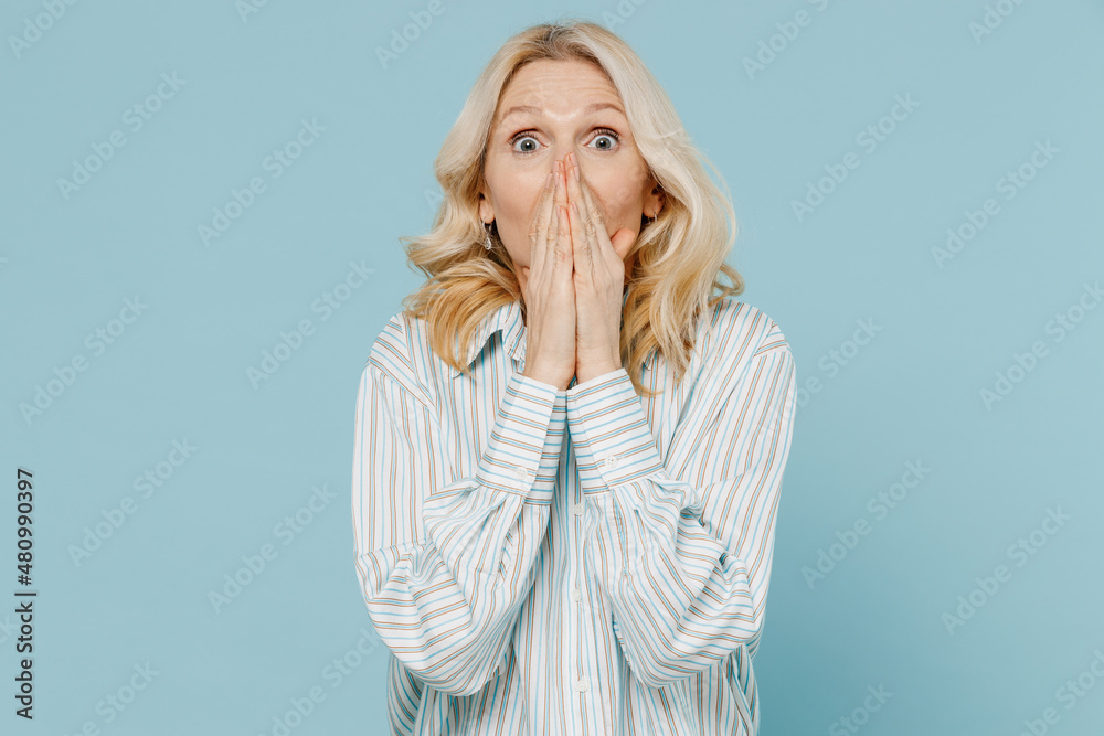 Elderly shocked surprised astonished caucasian woman 50s wear striped shirt cover mouth with hand look camera isolated on plain pastel light blue background studio portrait. People lifestyle concept.