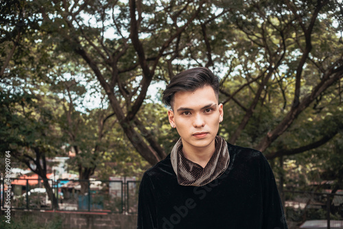 A handsome yet enigmatic young man with a straight-face looking at the camera. Outdoor scene at a park.