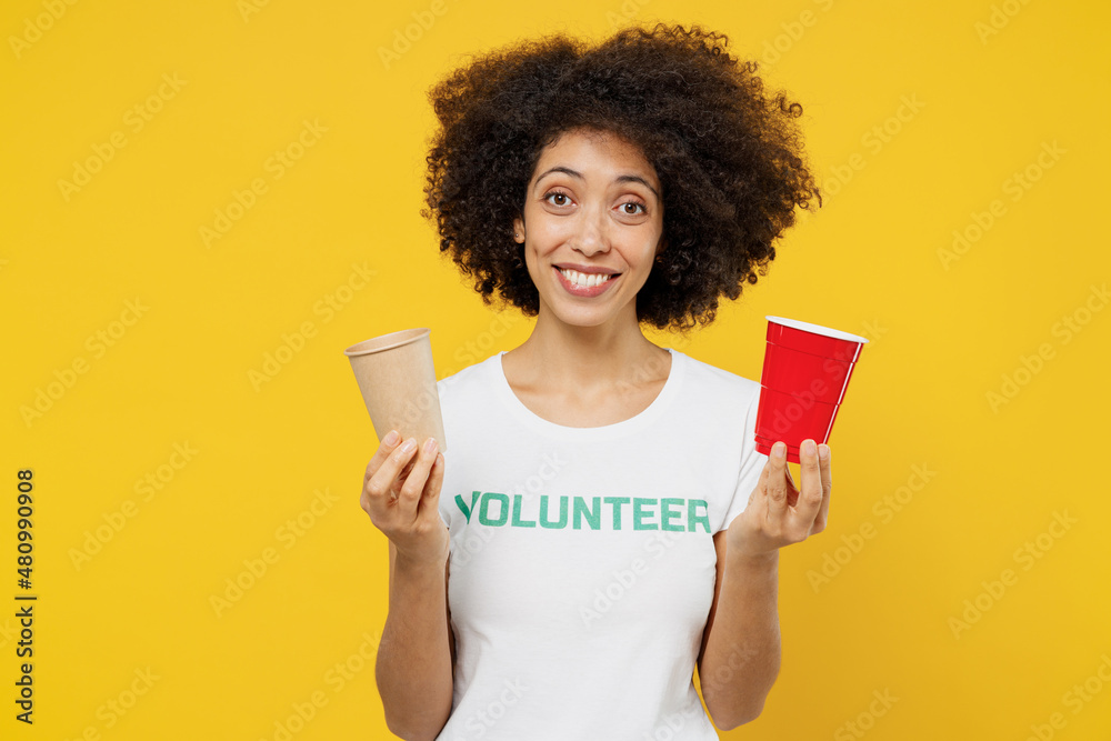 Young smiling woman of African American ethnicity in white volunteer tshirt hold choose paper plastic cup isolated on plain yellow background Voluntary free work assistance help charity grace concept Young smiling woman of African American ethnicity in white volunteer tshirt hold choose paper plastic cup isolated on plain yellow background Voluntary free work assistance help charity grace concept