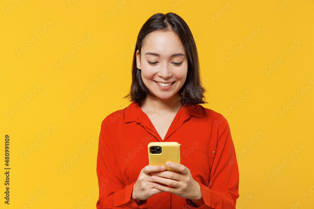 Charismatic vivid fun young woman of Asian ethnicity 20s years old wears orange shirt hold in hand use mobile cell phone reading searching chatting isolated on plain yellow background studio portrait. Charismatic vivid fun young woman of Asian ethnicity 20s years old wears orange shirt hold in hand use mobile cell phone reading searching chatting isolated on plain yellow background studio portrait.